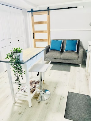 Una habitación con un escritorio blanco, un sofá gris con almohadas azules, una puerta de madera y un armario blanco.