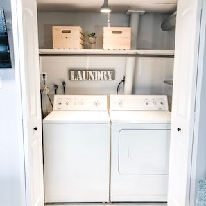 Una sala de lavandería con dos lavadoras blancas, un letrero de madera encima que dice "LAUNDRY" y una pequeña planta en maceta sobre la estantería.
