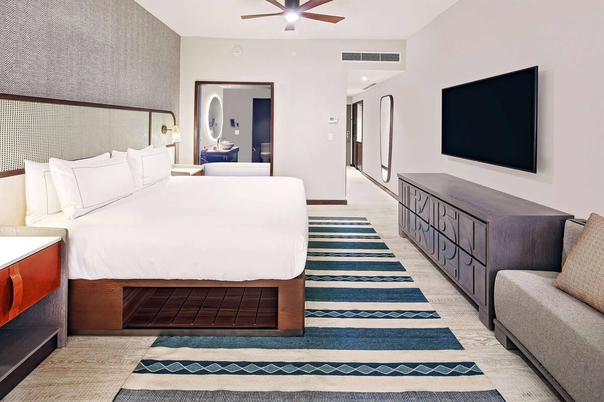 Ein Hotelzimmer mit einem großen Bett, einem Fernseher, einer Sitzecke, einem Spiegel und einem durchgegangenes Badezimmer.