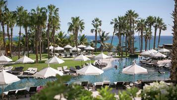 A resort pool area with lounge chairs and umbrellas, surrounded by palm trees and a view of the ocean.