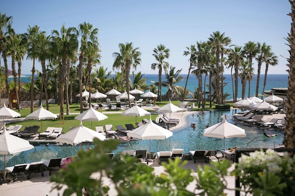 A resort pool area with lounge chairs and umbrellas, surrounded by palm trees and a view of the ocean.