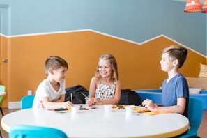 Three children sitting at a round table with art supplies, in a room with orange walls and blue chairs. - Hilton Grand Vacations Club La Pacifica Los Cabos (San José del Cabo)