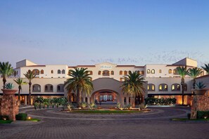 A large, multi-story hotel with a central arched entrance, palm trees, and a paved courtyard. - Hilton Grand Vacations Club La Pacifica Los Cabos (San José del Cabo)