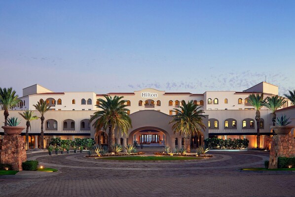 Un hotel grande de varios pisos con una entrada central en arco, palmeras y un patio empedrado.