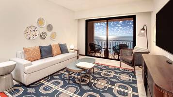 Un soggiorno moderno con un divano, un tavolino e un tappeto decorato. Si intravede un balcone con vista sul mare attraverso il vetro.