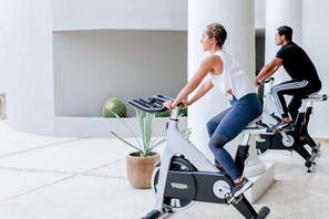 Dues persones pedalant en bicicletes estacionàries en un espai interior ben il·luminat, amb una planta en test i una columna blanca.