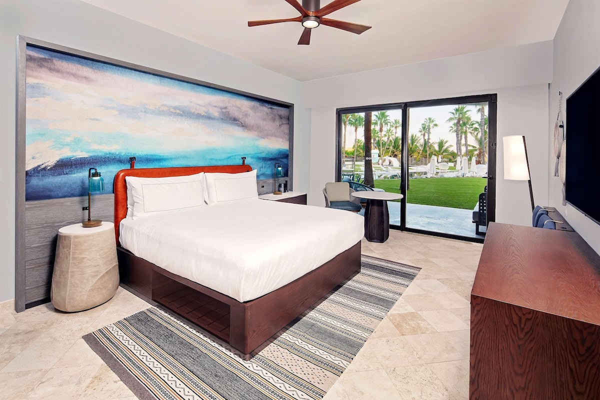 Une chambre d’hôtel moderne avec un grand lit, un ventilateur de plafond, un tableau représentant une plage, un bureau avec une lampe et une vue sur une piscine et des palmiers.