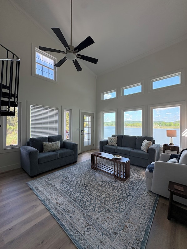 A spacious living room with a high ceiling fan, large windows, and a view of a body of water.
