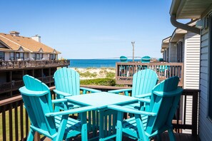 Un balcón con sillas Adirondack turquesas, una terraza de madera y vista a la playa y al océano.