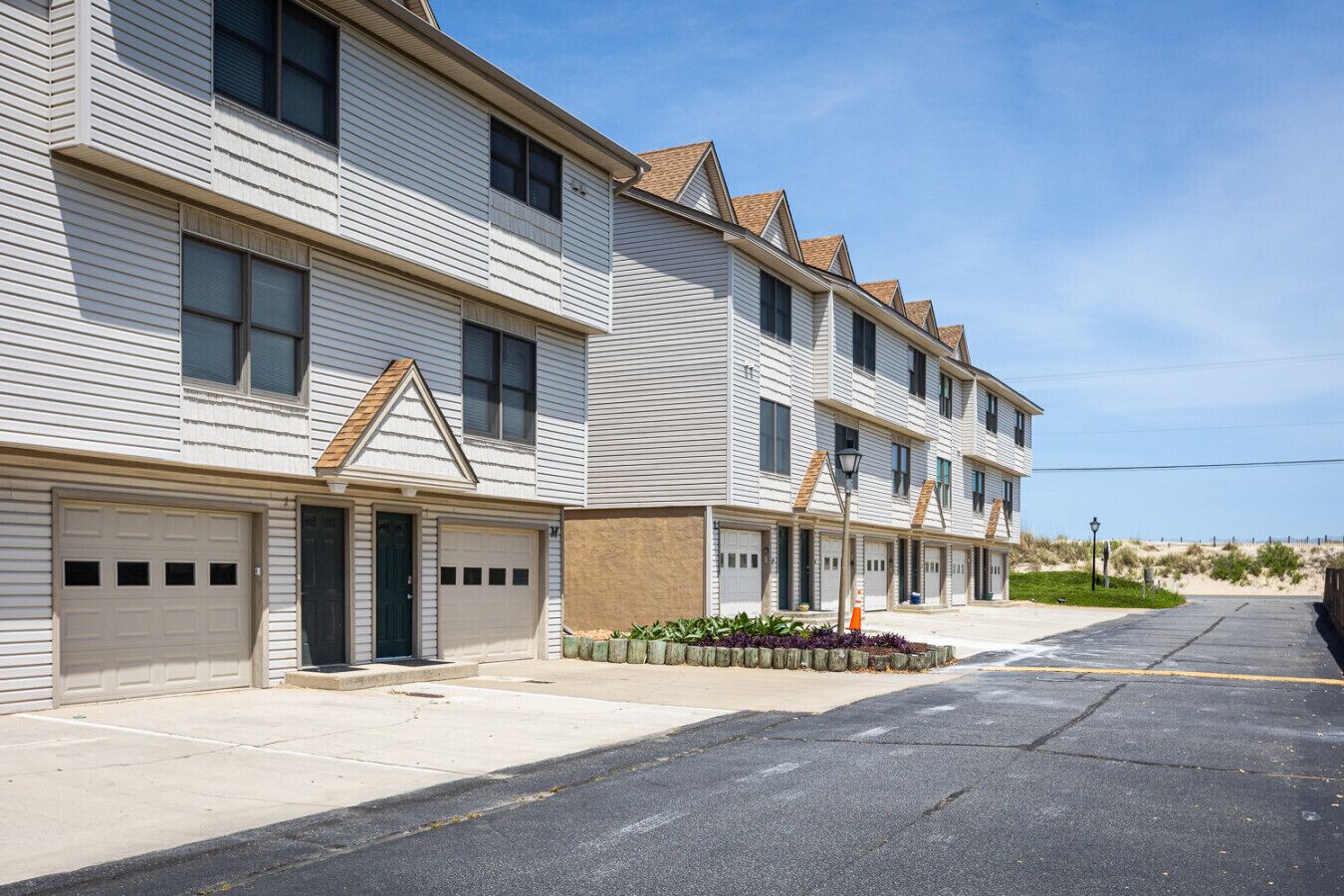A row of two-story townhouses with attached garages, a paved driveway, and a landscaped area with plants.
