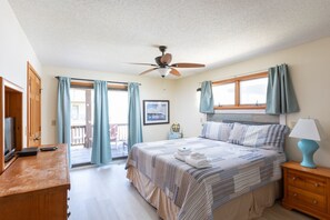A bedroom with a bed, a ceiling fan, a window with curtains, a bedside table with a lamp, and a view of the outdoors.