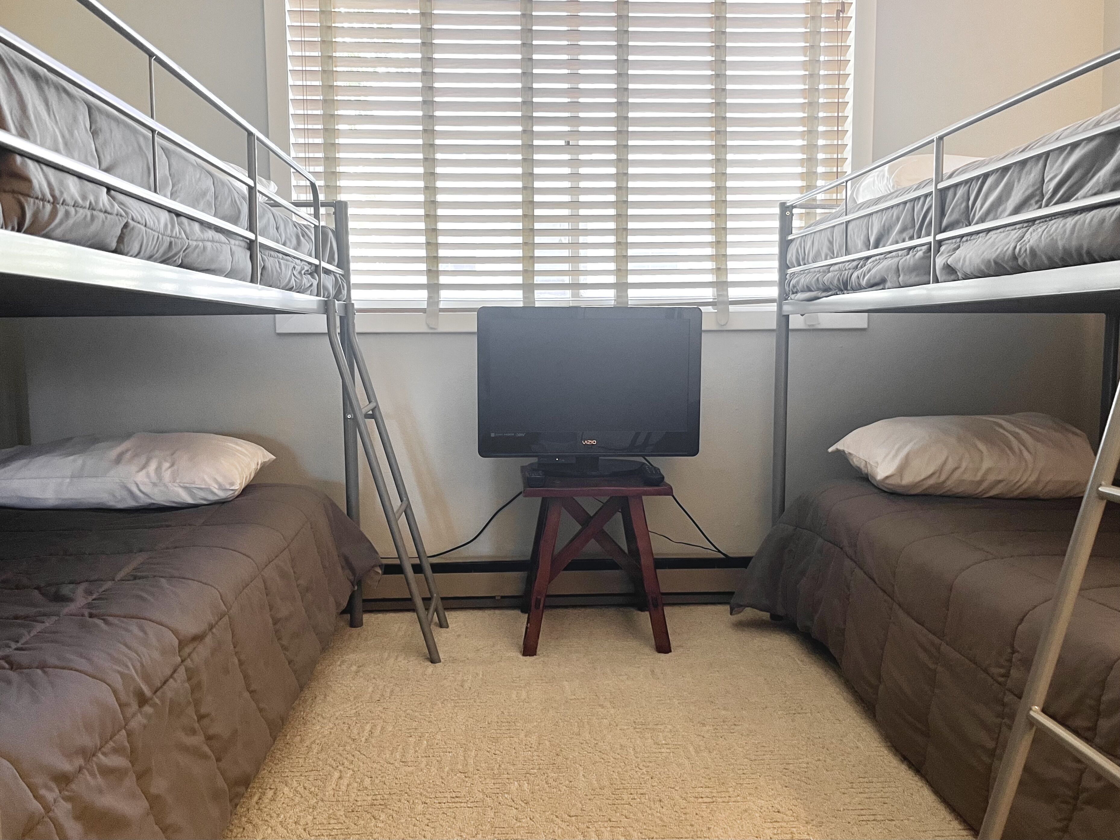 Une chambre de dortoir avec des lits superposés, un téléviseur posé sur un meuble en bois et une fenêtre avec des stores.