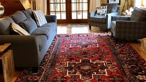Una sala de estar acogedora con techo de madera, un sofá gris, un sillón escocés, un tapete rojo estampado y una puerta con paneles de vidrio.