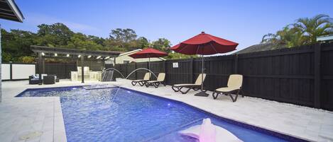 A swimming pool with a water feature, surrounded by lounge chairs and umbrellas, with a wooden fence and a covered patio area in the background.