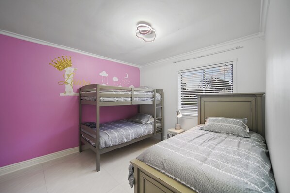 A bedroom with bunk beds, a pink wall with a crown and name decoration, a bedside table with a lamp, and a window with blinds.