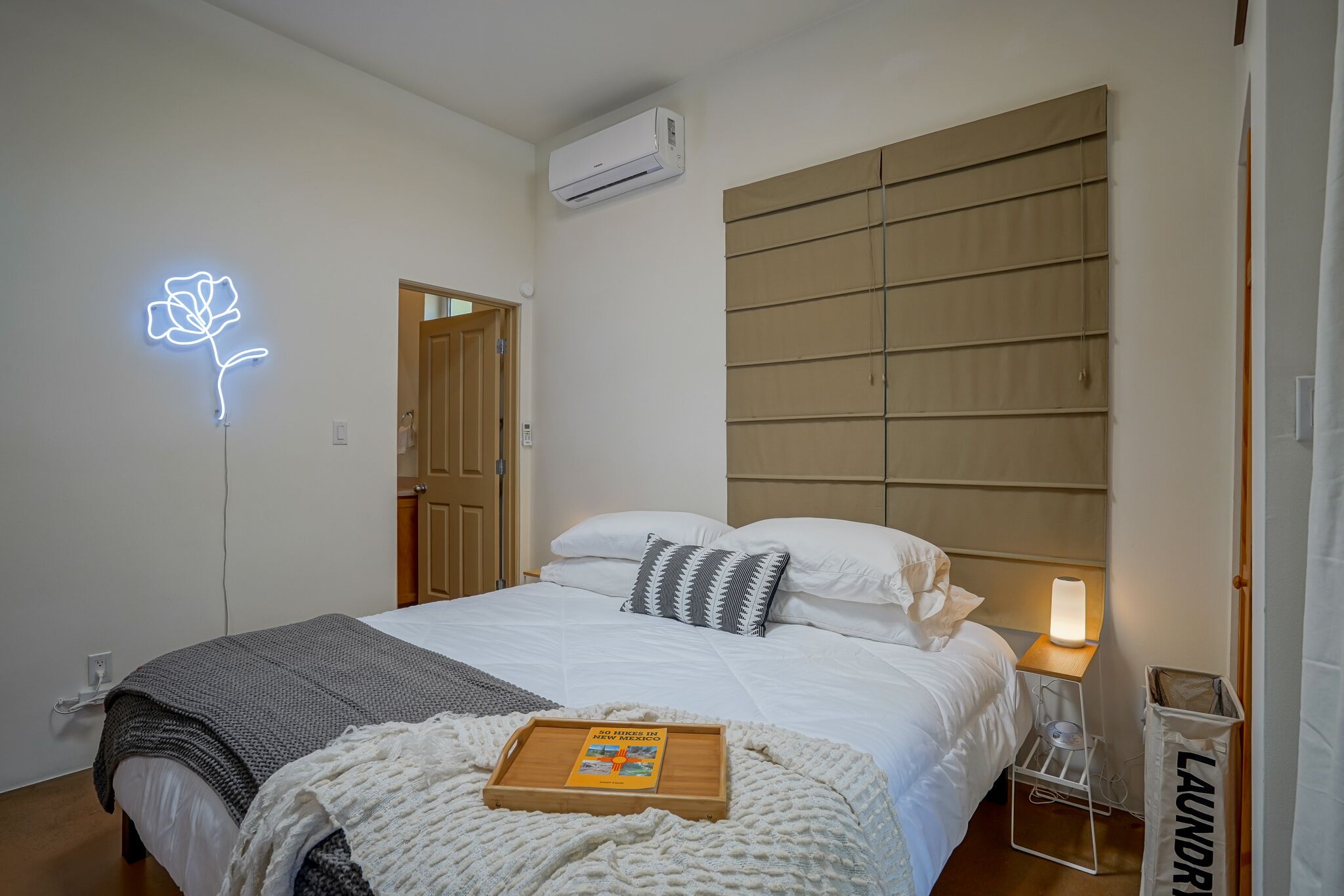 A neatly made bed with a bedside table, a wall-mounted air conditioner, and a neon rose sign.