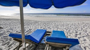 Ein Strand mit zwei blauen Liegestühlen unter einem blauen Sonnenschirm, der zum Meer hin ausgerichtet ist.
