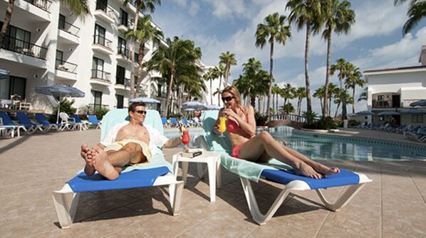 Two sun loungers by a poolside with palm trees and a hotel in the background.