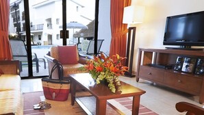 A hotel room with a sofa, a coffee table with a vase of flowers, a television on a wooden cabinet, and a view of the pool and buildings outside. - 2 Bedroom at The Royal Cancun  (Cancún)