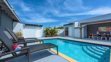 A swimming pool with lounge chairs and a table, surrounded by a white building with a tiled roof.