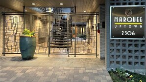 Entrada de hotel con una puerta metálica de diseño geométrico, una jardinera grande con plantas y un cartel que indica el nombre y la ubicación del hotel.