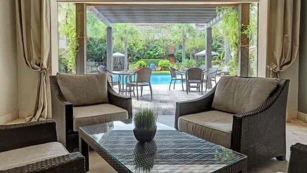 Un patio con muebles de mimbre, una mesa de vidrio y vistas a una piscina y follaje.