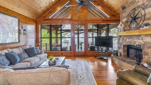 A spacious living room with a high ceiling, wooden beams, a stone fireplace, and a large TV. - Killen Time on Lake Martin (Dadeville)