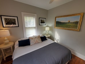 A bedroom with a bed, a nightstand with a lamp, a window with blinds, and two framed pictures on the wall.