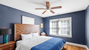 Een slaapkamer met een houten hoofdbord, een bed met wit en blauw beddengoed, een plafondventilator, een nachtkastje met een lamp en een raam met jaloezieën.