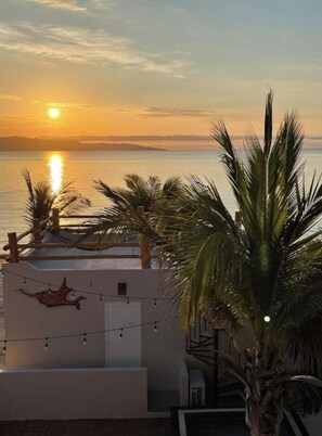 A sunset view from a balcony overlooking a body of water, with palm trees and a white building with a red decoration. - Beach Front Lofts 2 (el sargento)
