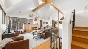 Een ruime woonkamer met een open haard, leren banken en een groot raam met uitzicht op een bos.