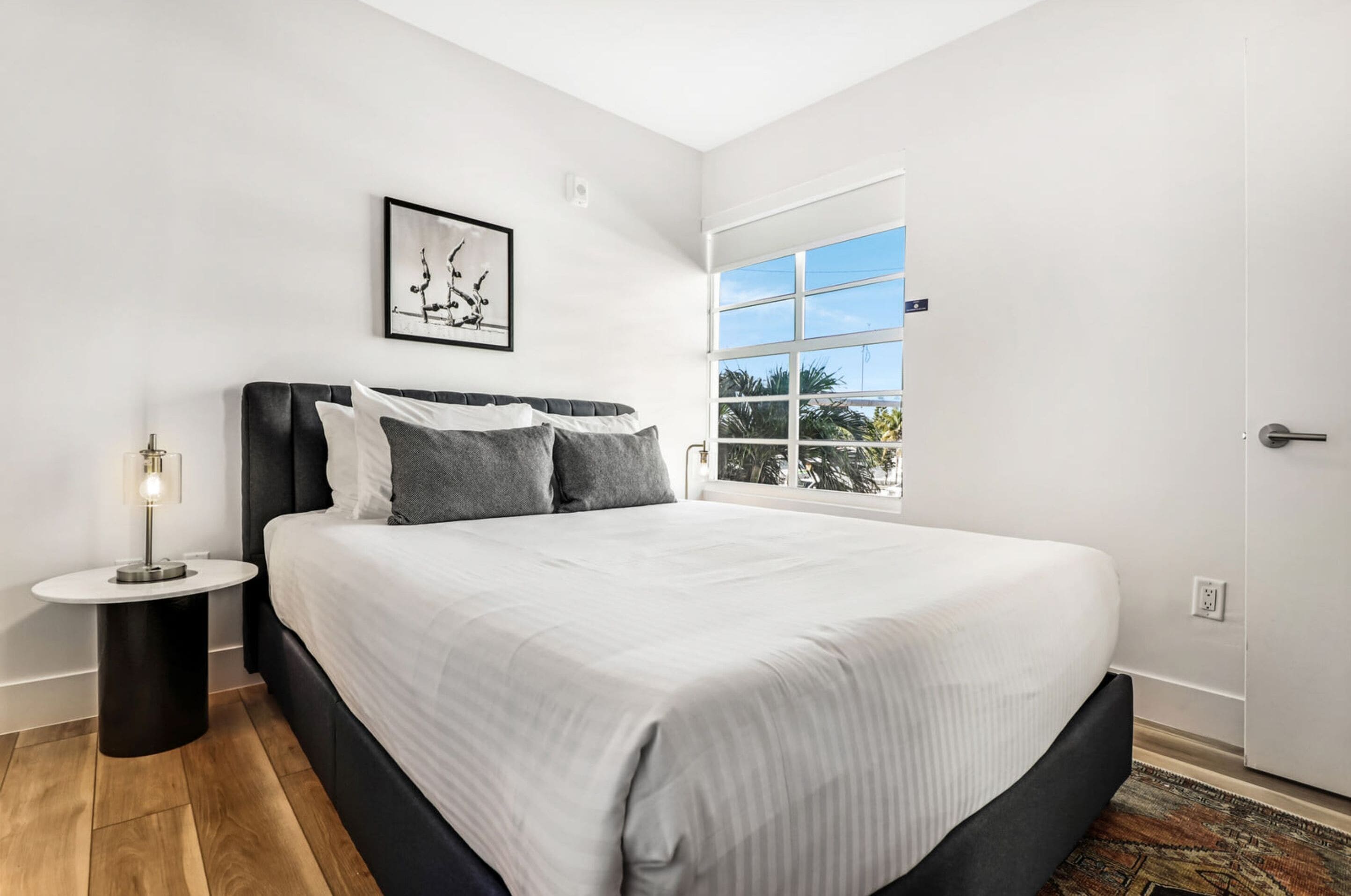 A modern bedroom with a large bed, a bedside table with a lamp, a framed artwork on the wall, and a window with a view of palm trees.