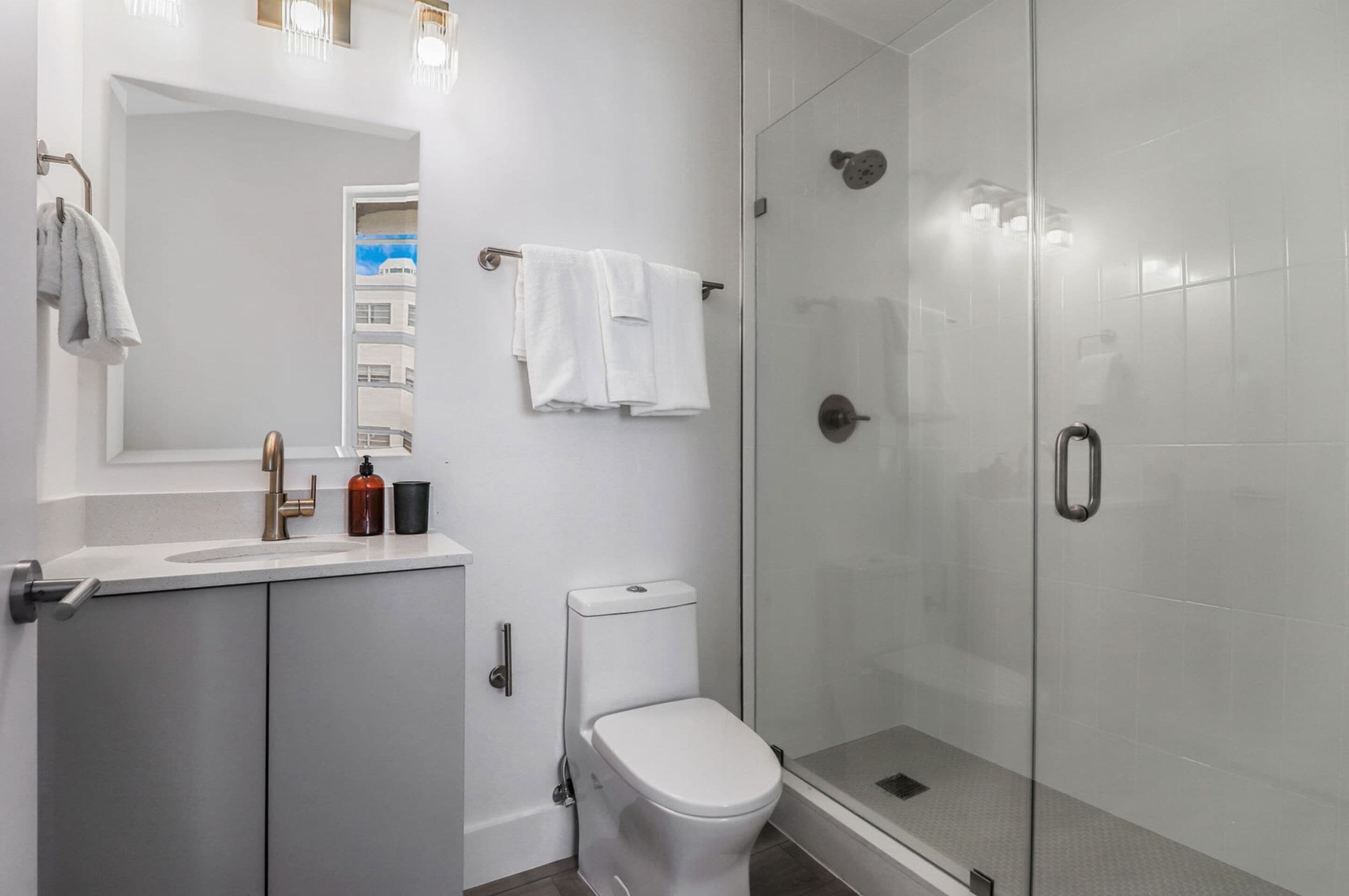 Une salle de bain moderne avec une douche à parois de verre, des toilettes blanches et un lavabo surmonté d’un miroir.