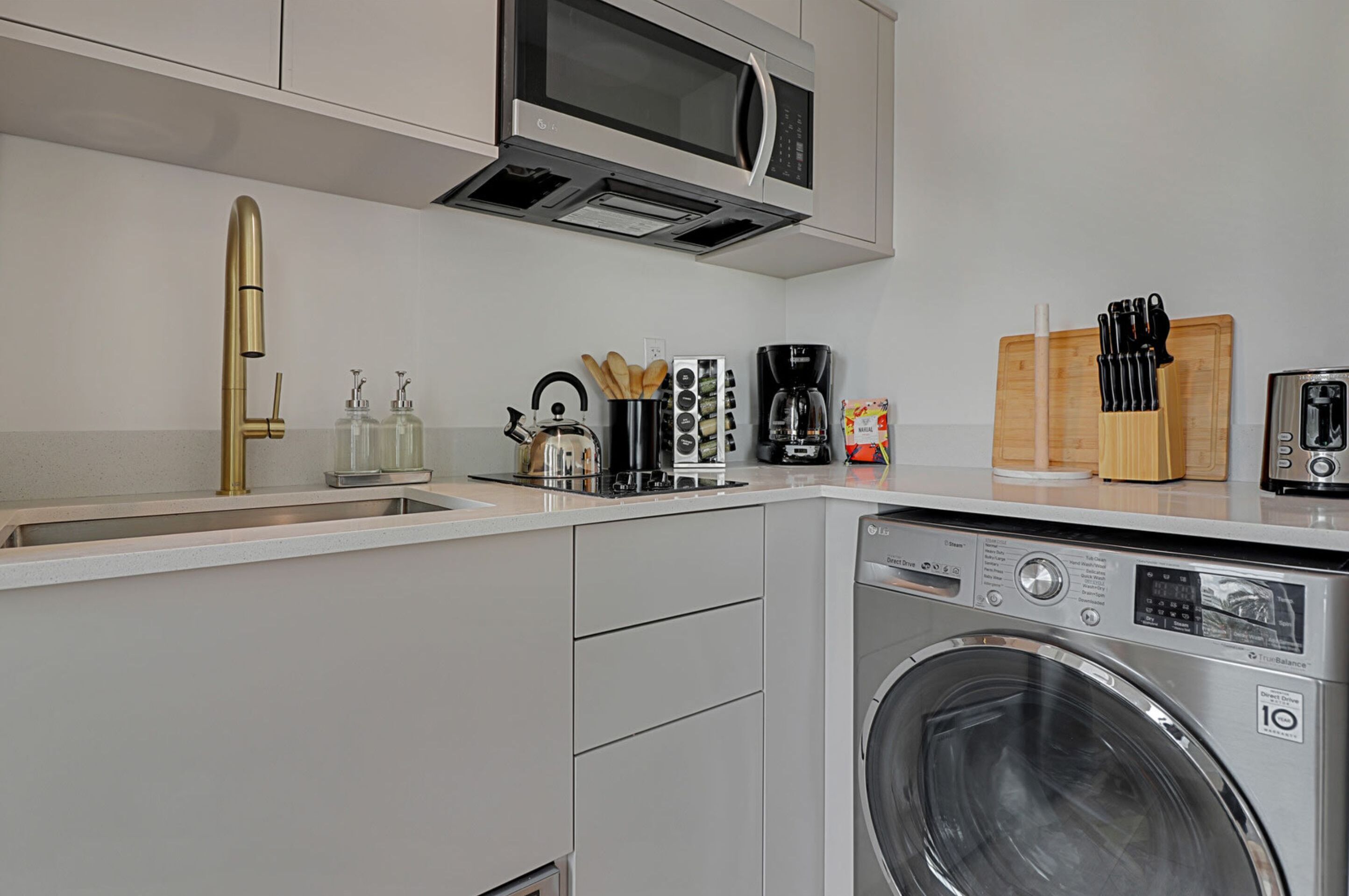 Een kleine keuken met een ingebouwde wasmachine, een magnetron, een spoelbak met een gouden kraan en een koffiezetapparaat.