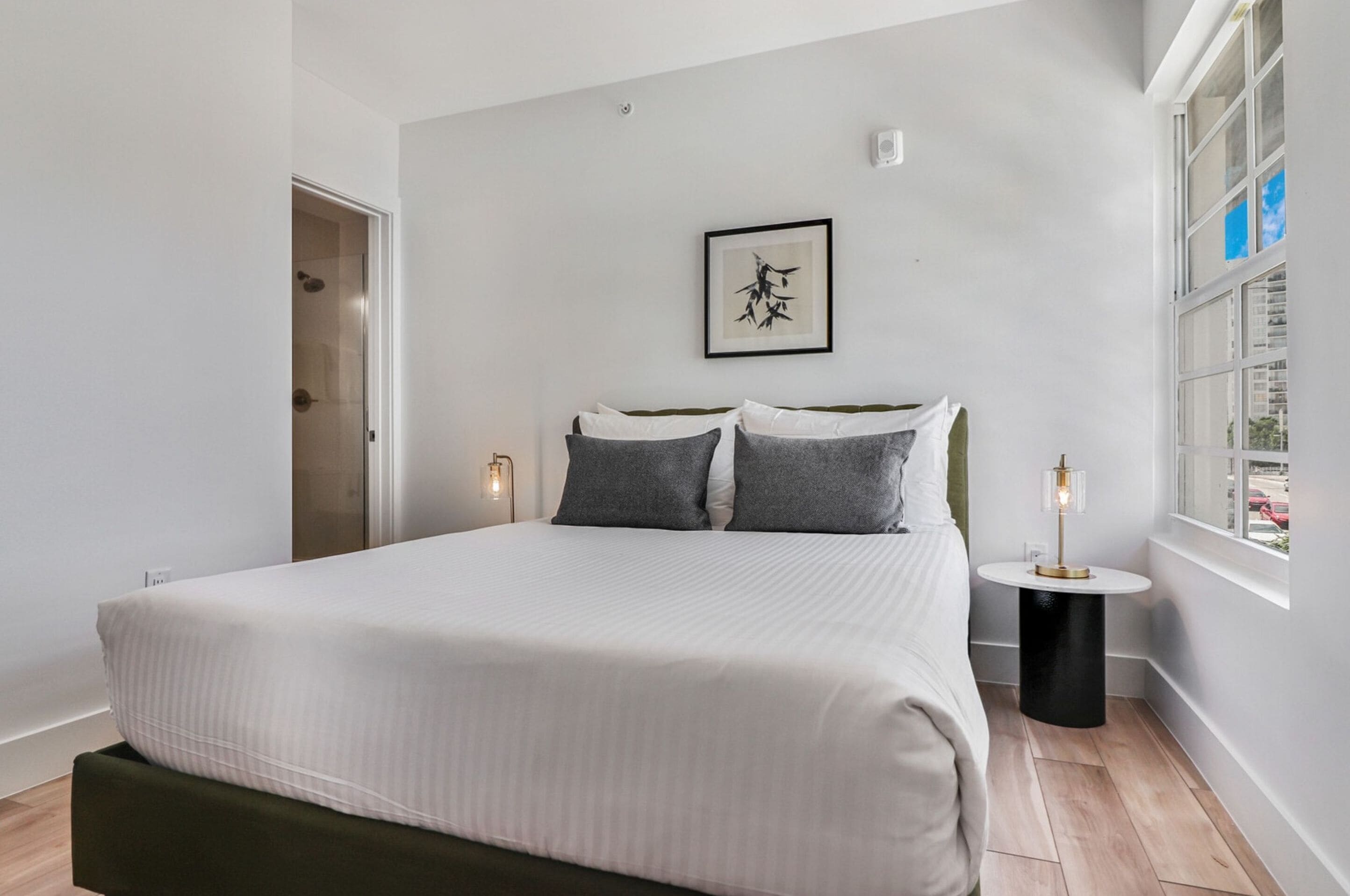 A bedroom with a bed, a bedside table, a window, and a framed artwork on the wall.
