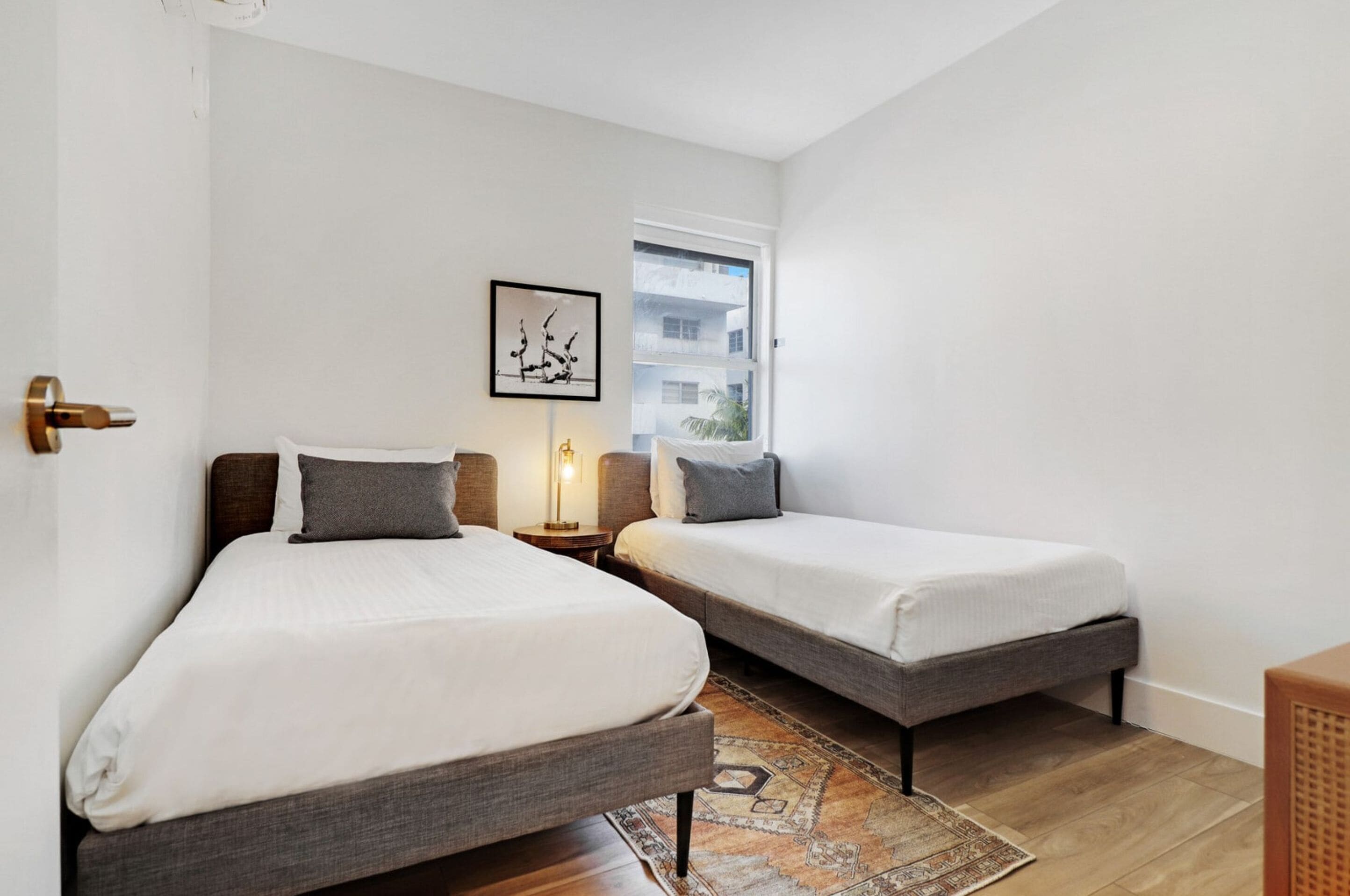 Une chambre d’hôtel avec deux lits simples, un petit tapis, une table de chevet avec une lampe et un tableau encadré au mur.