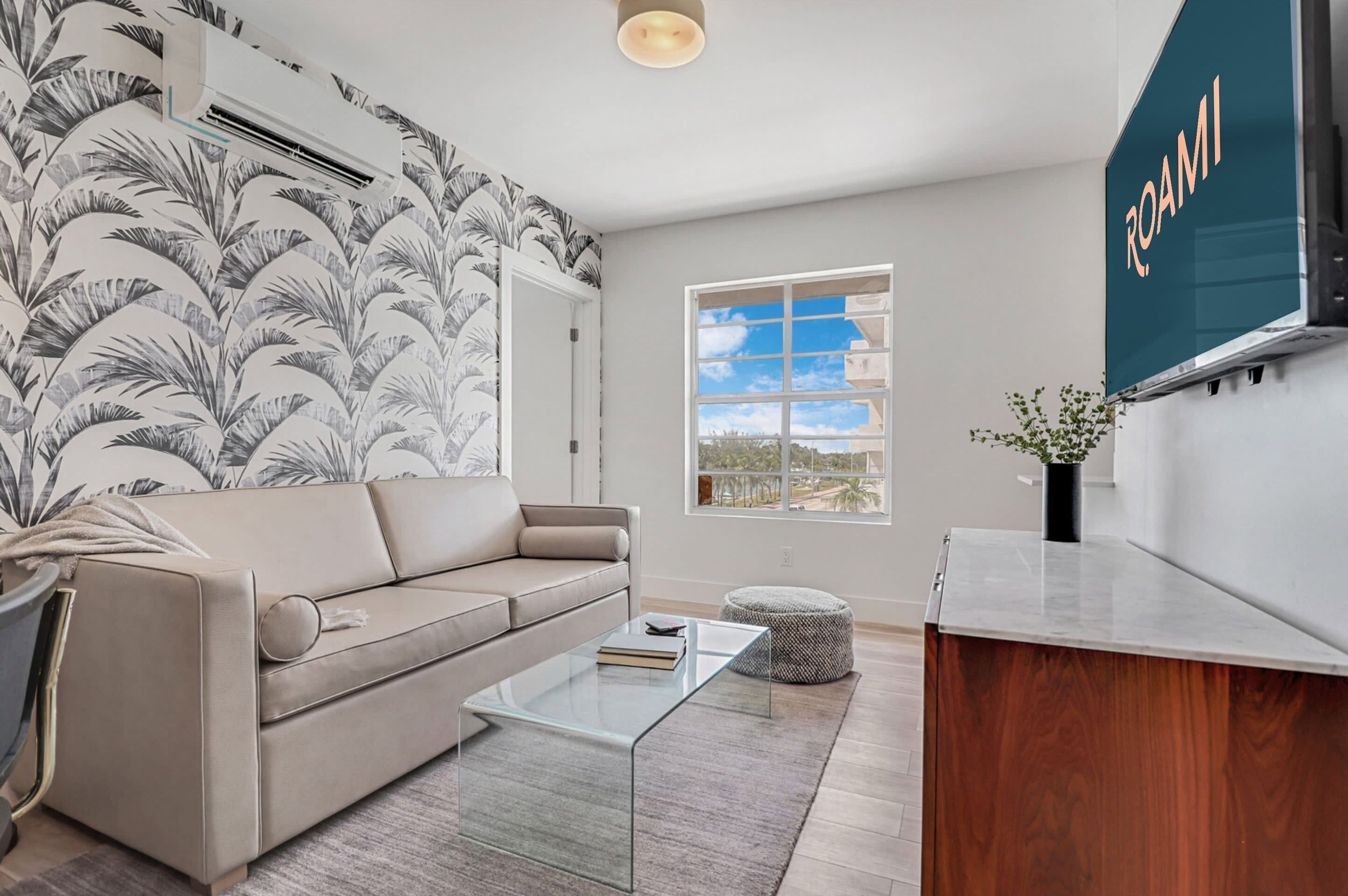 A modern living room with a beige sofa, a glass coffee table, a patterned wall, a window with a view, and a 'ROAMI' sign.