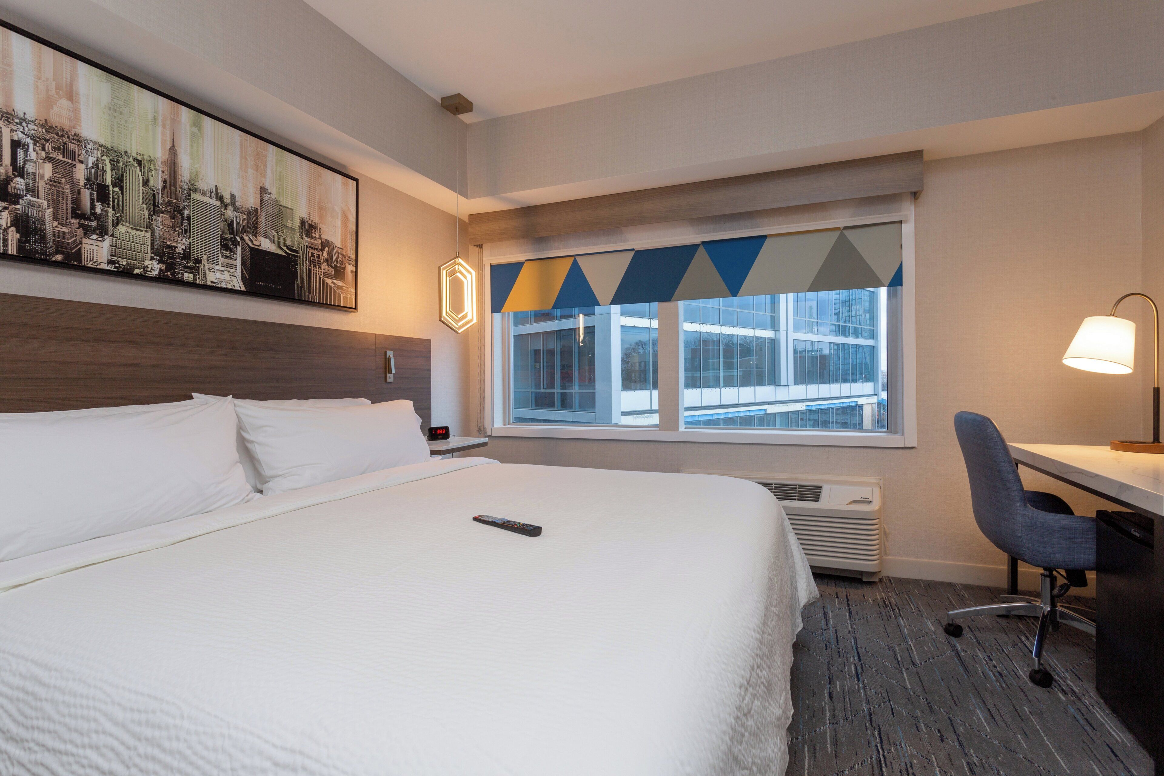 Een hotelkamer met een groot bed, een bureau met stoel, een raam met uitzicht op gebouwen en een schilderij aan de muur.