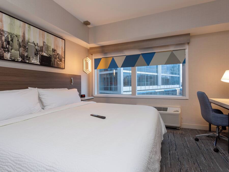 A hotel room with a large bed, a desk with a chair, a window with a view of buildings, and a painting on the wall.