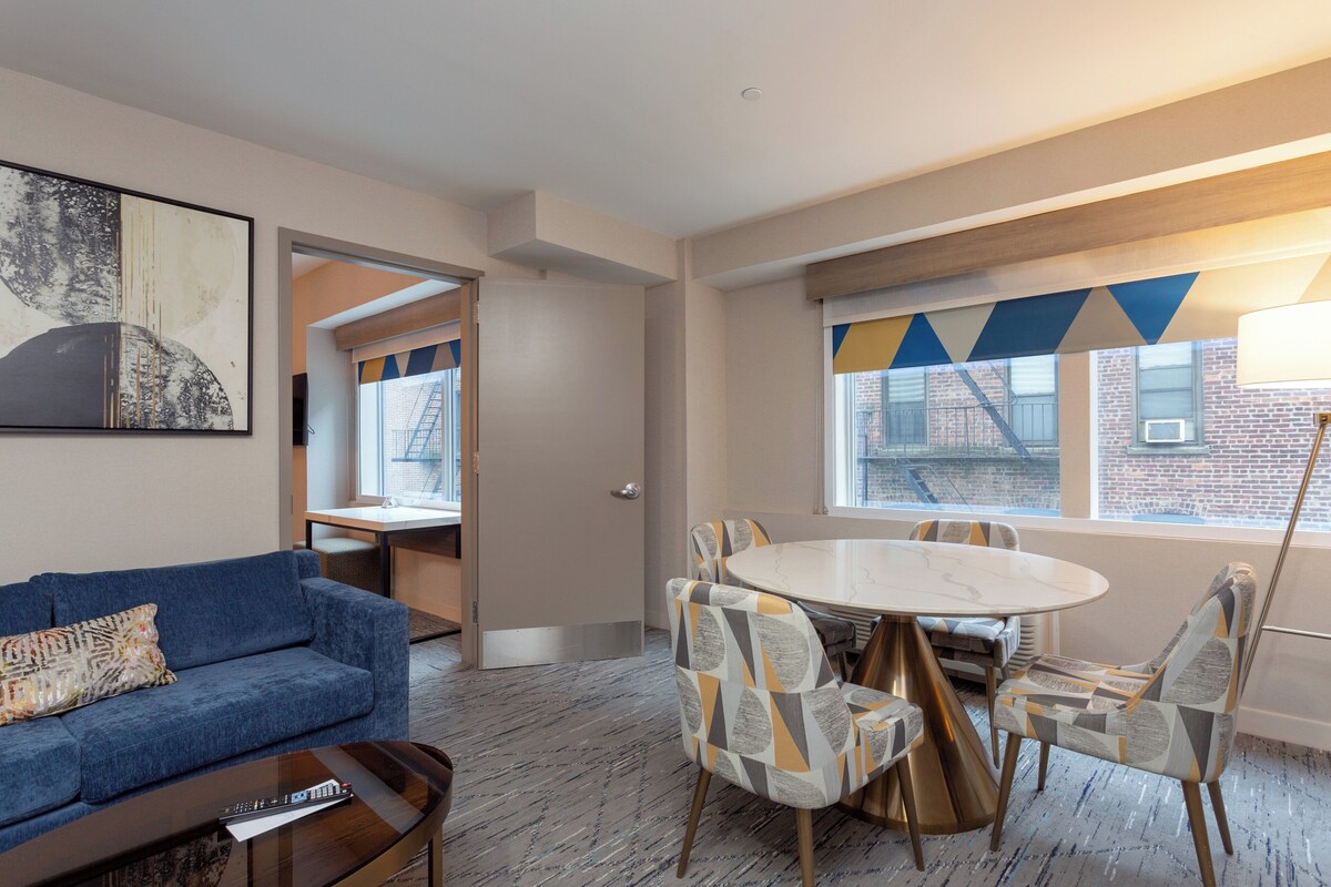 Une chambre d’hôtel moderne avec un canapé bleu, une table à manger ronde entourée de chaises à motifs et une grande fenêtre agrémentée d’un rideau à motifs géométriques.