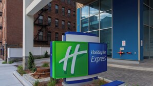 Une enseigne Holiday Inn Express aux couleurs verte et bleue, située devant un bâtiment moderne doté de grandes fenêtres et d’une façade en briques.