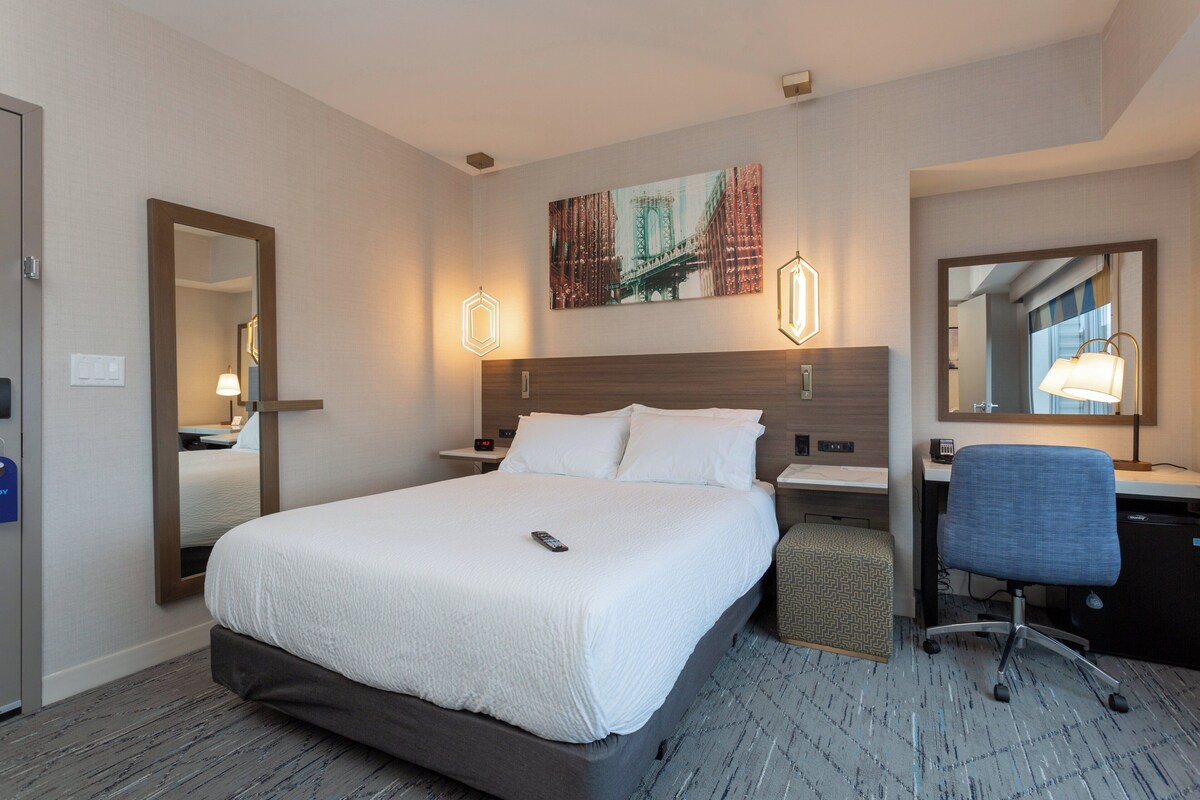 Une chambre d’hôtel avec un grand lit, un bureau avec une chaise, un miroir et un tableau représentant un pont accroché au mur.