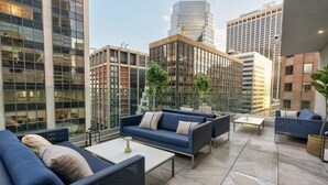 Un moderno salón en la azotea con sofás azules, una mesa de centro blanca y una barandilla de cristal con vistas a un paisaje urbano.
