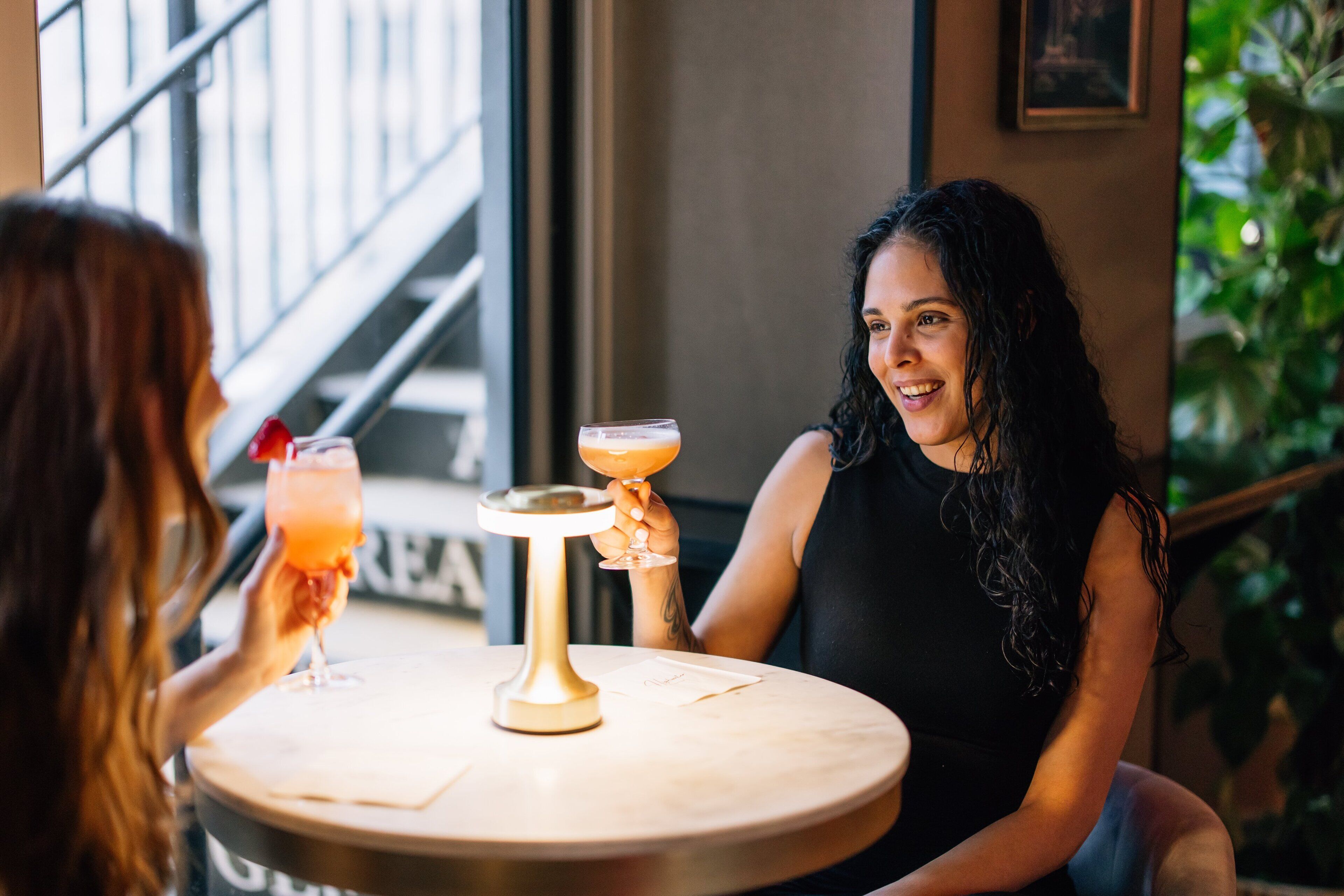 Twee personen zitten aan een ronde tafel met drankjes, een menukaart en een klein decoratief object.