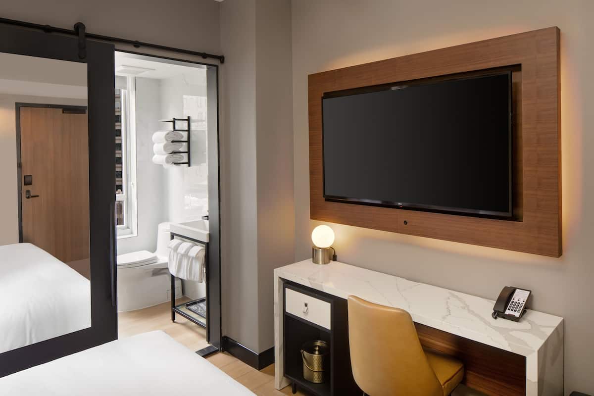 A hotel room with a bed, a desk with a phone, a TV mounted on the wall, and a bathroom visible through an open door.