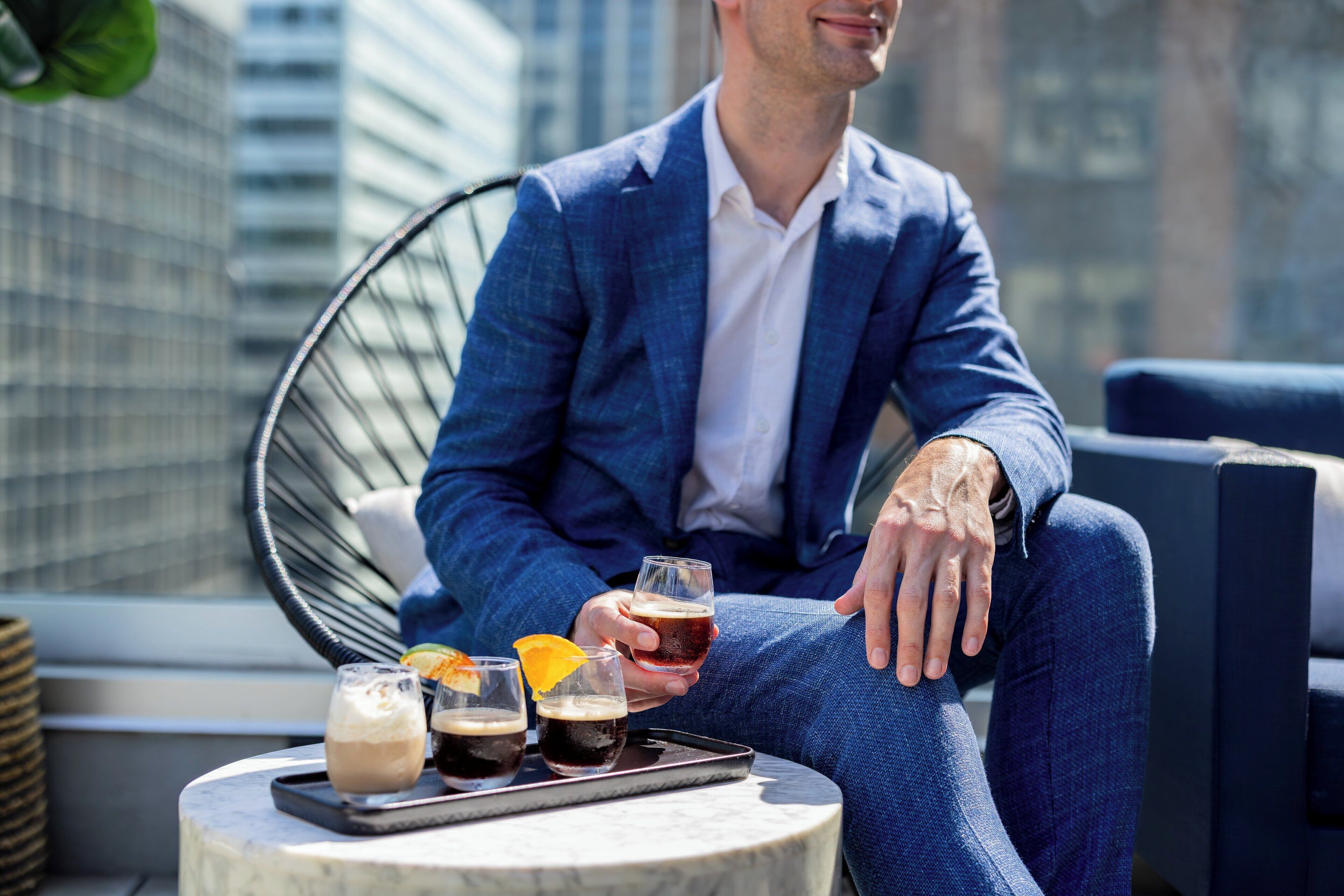 A man in a blue suit sitting on a chair with drinks on a table.