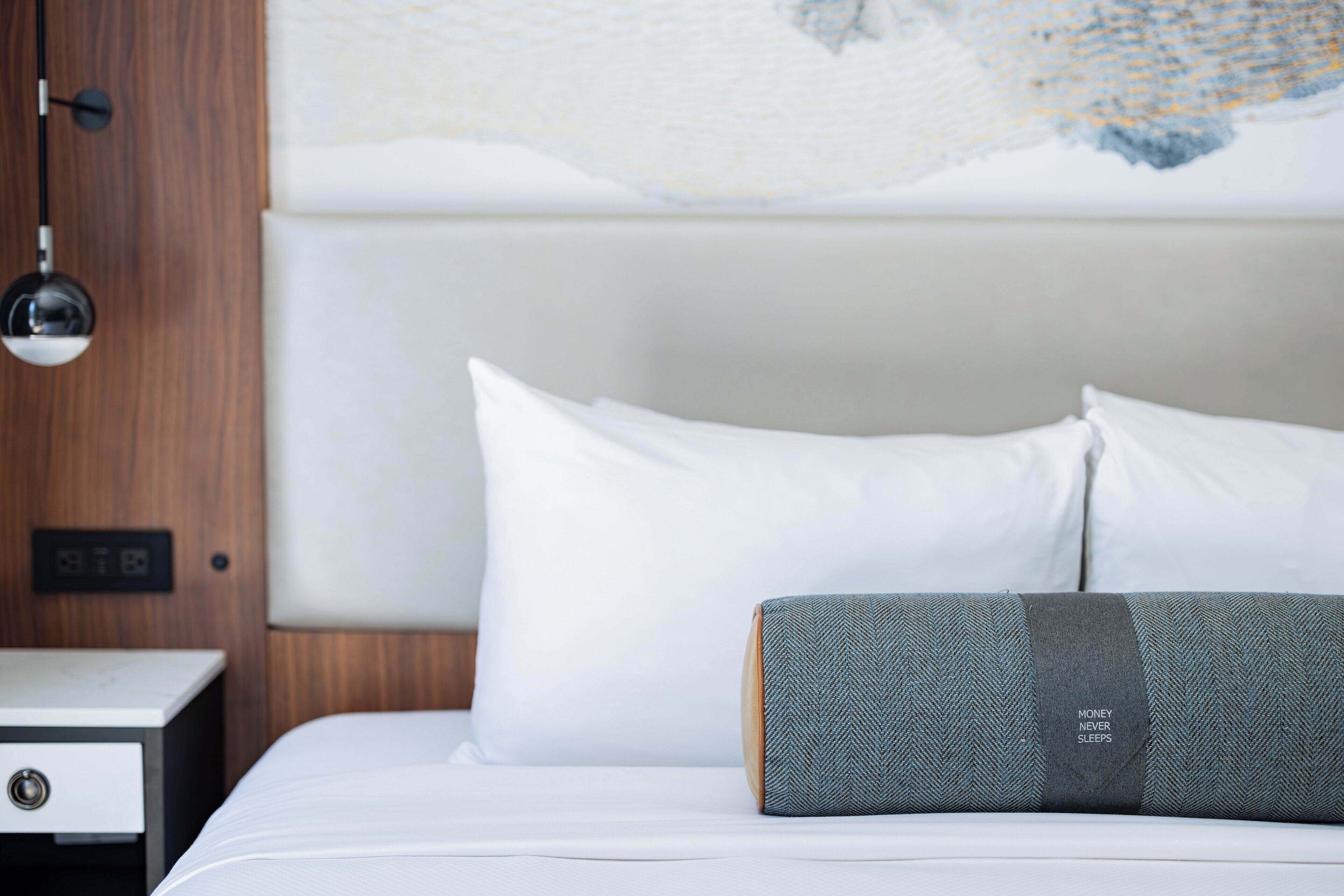 Una cama bien hecha con almohadas blancas y grises, una mesita de noche de madera con cajón, y una lámpara de pared.