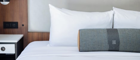Una cama bien hecha con almohadas blancas y grises, una mesita de noche de madera con cajón, y una lámpara de pared.