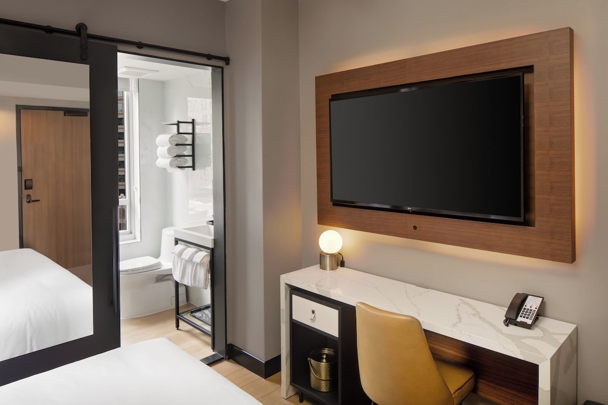 Habitación de hotel con cama, escritorio con teléfono, televisor montado en la pared y baño visible a través de una puerta abierta.