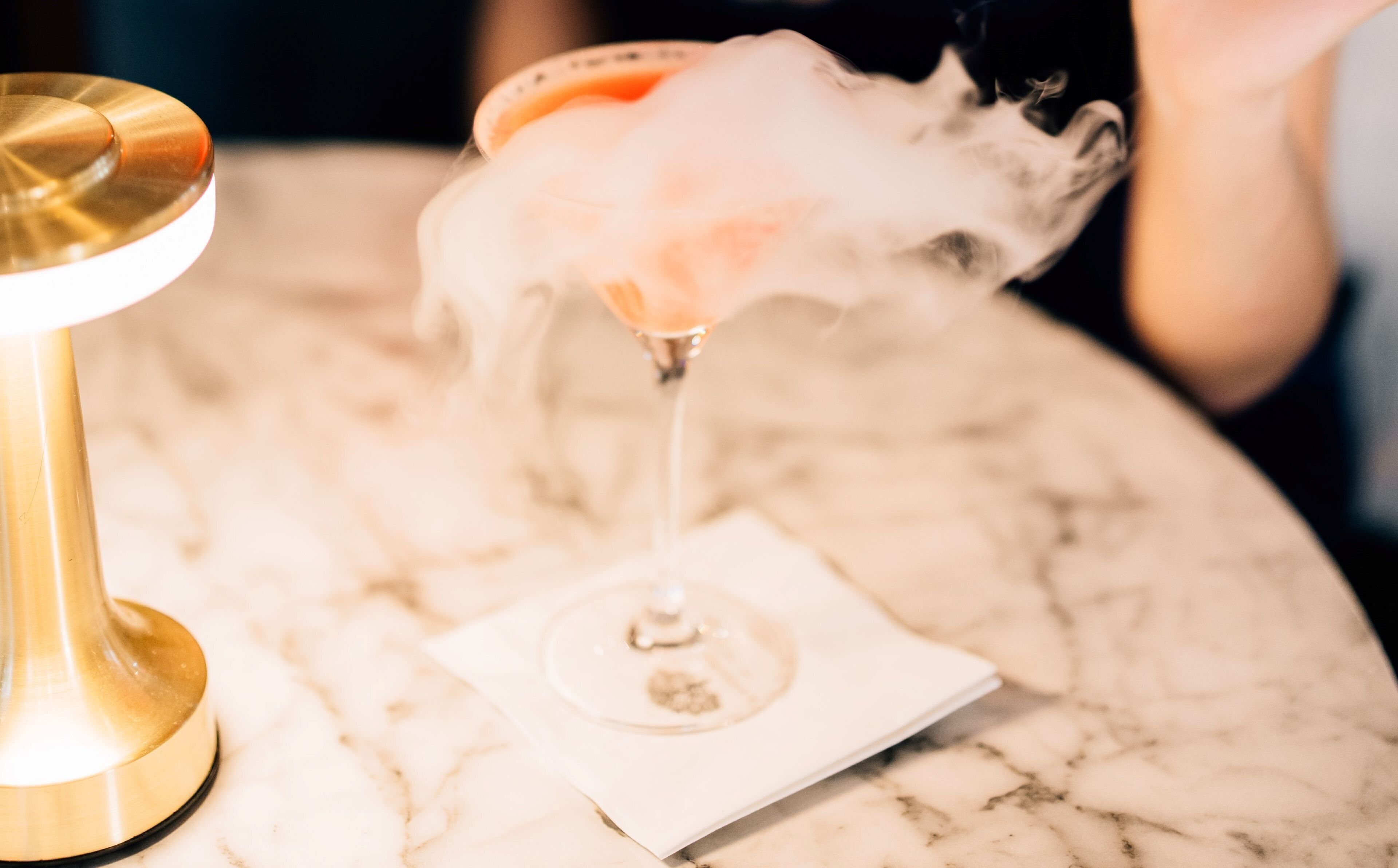 A cocktail glass with smoke, a gold-colored base, and a white napkin on a marble surface.